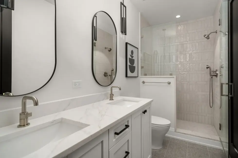 Double vanity with white drawer fronts, a stone-look top, brushed-nickel faucets and oval black-framed mirrors, glass shower beyond