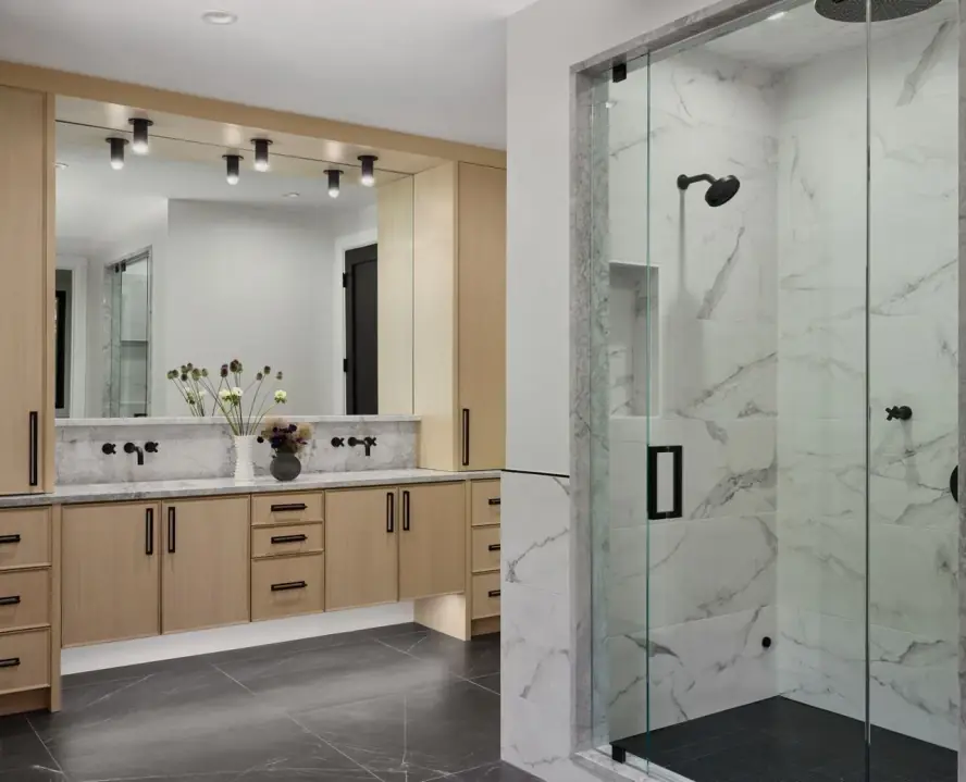 Primary bathroom with an oak double vanity, marble-look walls and a glass shower