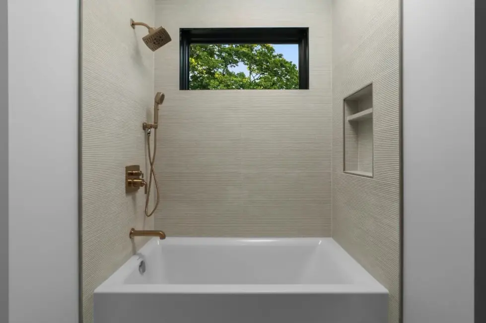 Alcove tub with ribbed light tile walls, a brushed brass shower set, a recessed niche and a window