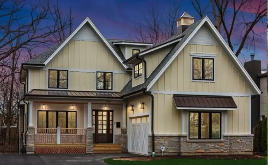 Cream board-and-batten home at dusk with a stone base and a standing-seam metal porch roof