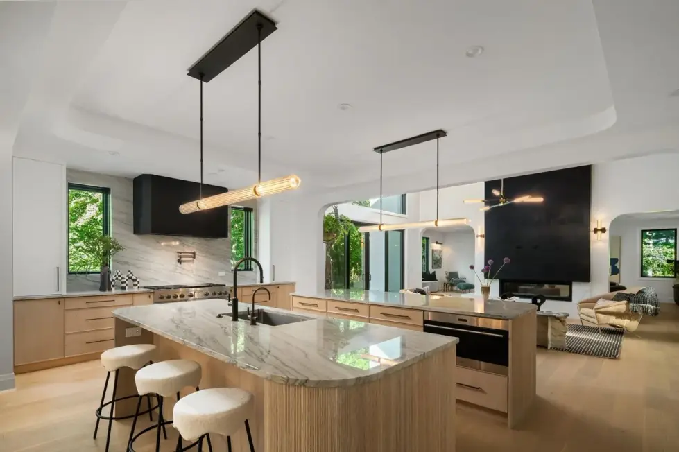 Open kitchen with two stone-topped oak islands, a black range hood, linear pendants and a view into the family room